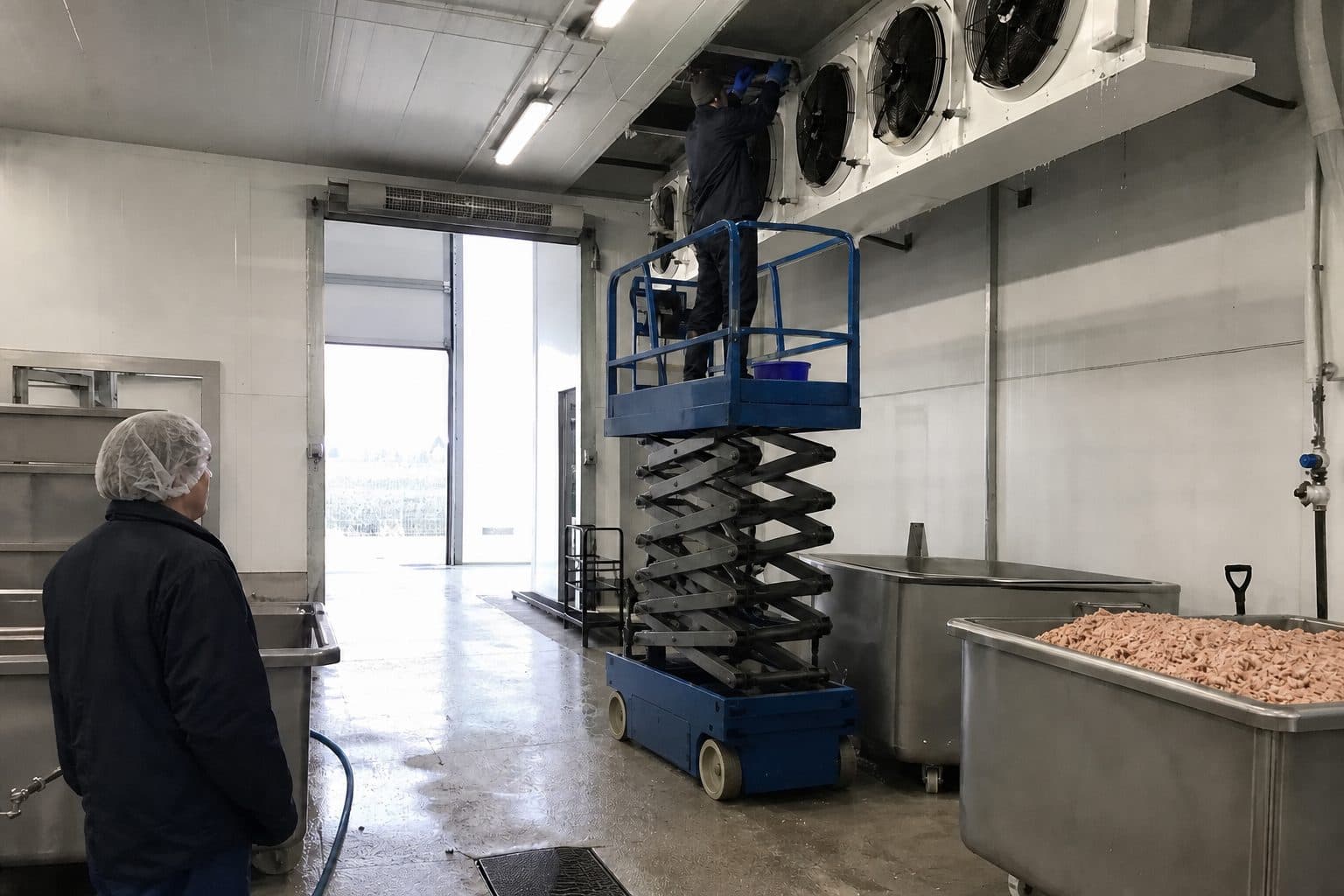 A technician on a lift servicing a ceiling-mounted evaporator in a food processing plant cold room while stacked pallets stand below