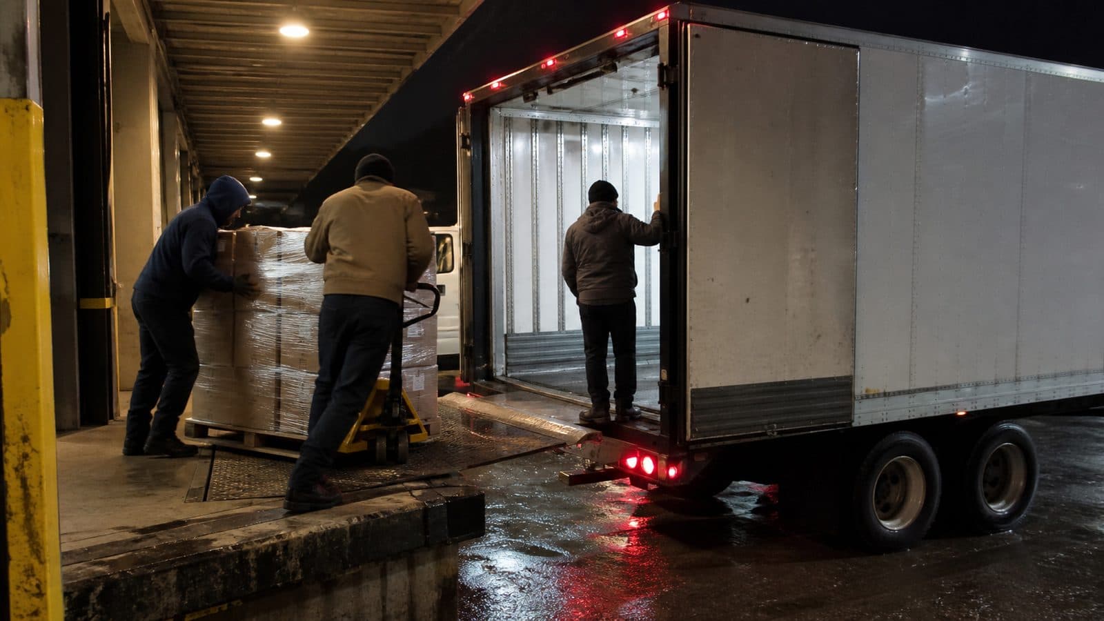 Warehouse staff moving pallets from a cold room into a refrigerated trailer backed to the dock at night