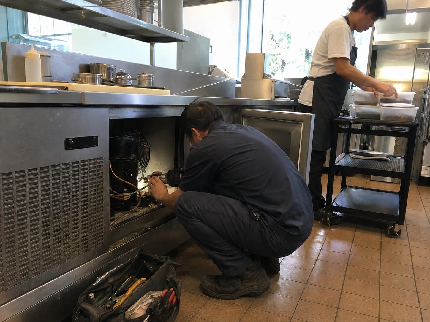 A technician working on the compressor of a refrigerated prep-line unit in a commercial kitchen while the kitchen stays in operation